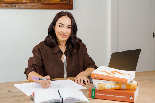 Persona tecleando en un ordenador, con un libro abierto al lado, un café y una pila de libros detrás del portátil